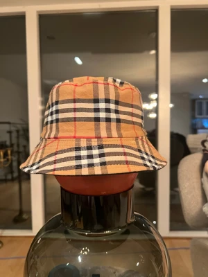 Burberry beige bucket hat - Säljer en klassisk bucket hat från Burberry i beige med det ikoniska rutmönstret i svart, vitt och rött. Hatten är tillverkad i bomull och har en avslappnad passform. Perfekt accessoar för att lyfta din streetwear-look.
