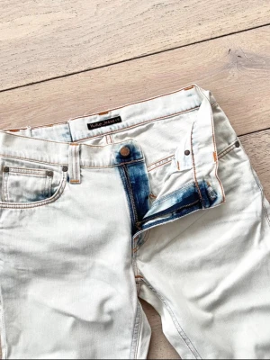 Bleached jeans från Nudie Jeans - Säljer ett par ljusbeiga raka jeans från Nudie Jeans med klassisk femficksdesign och kontrastsömmar. Jeansen har en unik blå detalj vid gylfen och är tillverkade i denim. Perfekta för en avslappnad och trendig look.   W30 l34