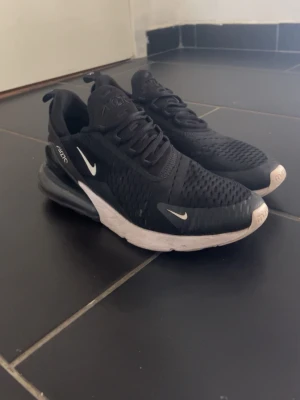 Nike Air Max 270 svarta sneakers - Svarta Nike Air Max 270 sneakers med vit sula och synlig Air-enhet i hälen. Ovandelen är i mesh för bra ventilation och har vita Nike-loggor på sidan och tungan. Snygg och sportig design som passar till streetwear.