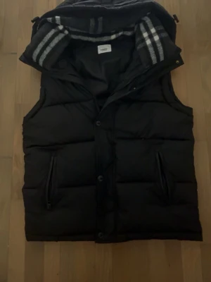 Burberry väst  - Snygg svart dunväst från Burberry med avtagbar huva och klassiskt rutigt foder. Västen har två dragkedjeförsedda fickor och knäppning framtill. Perfekt för lager-på-lager och håller dig varm under kyliga dagar.