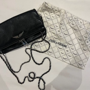 Svart axelväska från Zadig & Voltaire - Snygg svart axelväska i skinn från Zadig & Voltaire med kedjerem och coolt vingmotiv. Väskan har dragkedja och kommer med en praktisk dustbag. Perfekt för att ge din outfit en edgy touch!