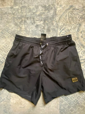 Versace badshorts - Snygga svarta badbyxor från Versace med elastisk midja och dragsko. Innerbyxa i mesh för extra komfort och en liten gulddetalj med logga på benet. Perfekta för stranden eller poolen och ger en lyxig känsla.