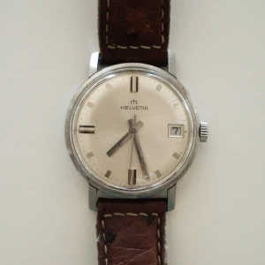 Vintage Helvetia | Strutsläder - Vintage armbandsur från Helvetia med klassisk schweizisk design. Mekaniskt manual-wind urverk med datumvisning. Boett i stål och ljus originaltavla med indexmarkeringar. Klockan har naturlig vintage-patina och normalt bruksslitage som hör till åldern. Monterad på brunt armband i strutsläder. Säljs i befintligt skick och är inte servad. Perfekt för dig som uppskattar klassiska schweiziska klockor från 1960-talet.