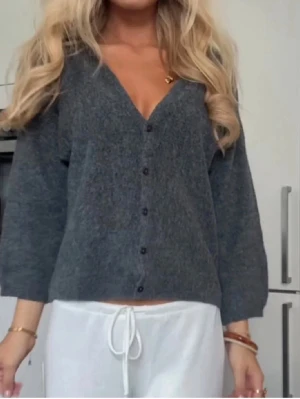 Mörkgrå kofta med knappar - Mysig mörkgrå kofta med v-ringning och knappar framtill. Koftan har långa ärmar och en avslappnad passform, perfekt att slänga på sig över ett linne eller t-shirt. Materialet ser ut att vara stickat och mjukt.