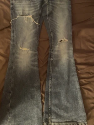 Blå slitna bootcut jeans Gina Tricot  - Säljer ett par blå bootcut lågmidiade jeans från Gina Tricot med slitningar och hål på låren och knäna. Jeansen har en klassisk femficksmodell och är tillverkade i denim med tvättad look. Perfekta för en avslappnad och trendig stil.