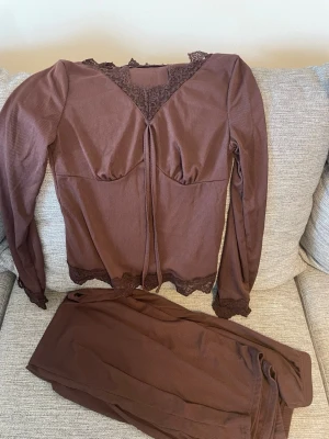 Brun pyjamas med spetsdetaljer - Mysig brun pyjamas i två delar med långärmad topp och långa byxor. Setet har fina spetsdetaljer vid halsringning, ärmslut och midja. Materialet är mjukt och ribbat för extra komfort. Perfekt för avslappnade kvällar hemma. Aldrig använd 