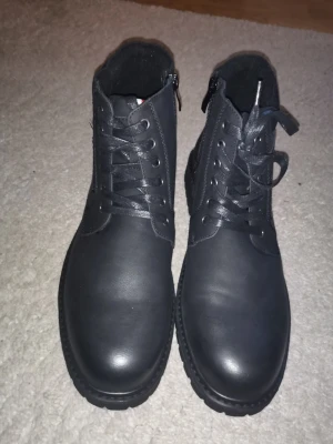 Svarta kängor med dragkedja - Snygga svarta kängor i matt skinnimitation med snörning och dragkedja på sidan. Grov sula för extra grepp och metallfärgade öljetter. Perfekta för dig som vill ha en stilren och robust look till hösten eller vintern.