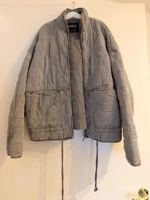 Grå quiltad cardiganjacka från Zara - Quiltad grå cardiganjacka från Zara med stora fickor framtill och dragsko i nederkant. Jackan har ett vågigt mönster i tyget och hög krage. Perfekt för lager-på-lager och en chill look. Tvättar absolut jackan innan eftersom de finns en fläck men den går bort🤍