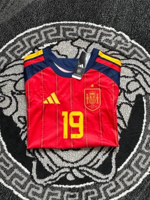 Spaniens matchtröja Adidas röd - Säljer en officiell röd matchtröja från Spaniens landslag med nummer 19. Tröjan har gula detaljer, blå ärmar och tunna vertikala ränder. Adidas-logga på bröstet och landslagets emblem i gult. Tillverkad i lätt funktionsmaterial, perfekt för fotboll.