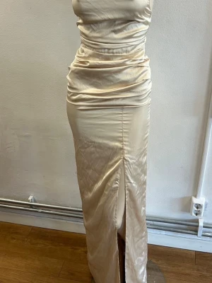 Champagnefärgad långklänning med slits - Elegant playlist långklänning i champagnefärg med smala axelband som korsas i ryggen och snörning baktill. Klänningen har en slits framtill och är tillverkad i ett glansigt satinliknande material som ger en lyxig känsla. Storlek S. 