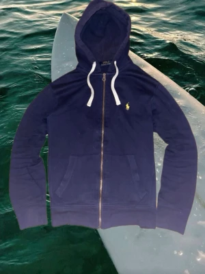 Mörkblå hoodie från Polo Ralph Lauren - Snygg mörkblå hoodie från Polo Ralph Lauren med dragkedja framtill och vita band i huvan. Klassisk gul logga broderad på bröstet.       Perfekt för en avslappnad och stilren look.                Längd: 63cm.                                                                 Bredd: 44cm