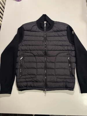 Moncler cardigan - Knappt använd Moncler cardigan, passar dig som runt 175-185, ser oanvänd ut. Snygg mörkblå pufferjacka från Moncler med stickade ärmar och quiltad front. Jackan har två dragkedjefickor framtill, hög krage och Moncler-logga på ärmen. Perfekt för kyliga dagar och ger en sportig men stilren look. 