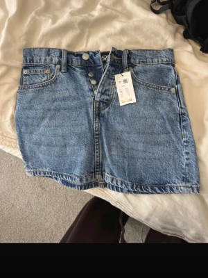 Blå jeanskjol med knappar - Snygg blå jeanskjol i klassisk modell med knappar framtill och femficksdesign. Kjolen har en kort längd och är tillverkad i slitstarkt denimtyg. Perfekt att matcha med en enkel t-shirt eller hoodie för en avslappnad look.