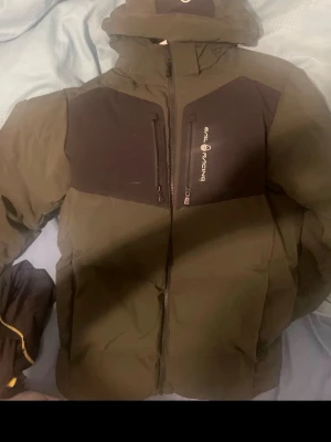 Grön dunjacka från Sail Racing - Snygg grön dunjacka från Sail Racing med svarta partier över bröstet och två vertikala bröstfickor med dragkedja. Jackan har huva och logga på bröstet. Perfekt för kalla vinterdagar och riktigt varm tack vare dunfyllningen.