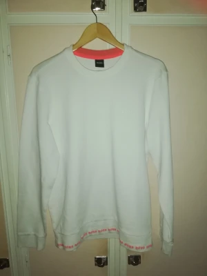 Hugo Boss sweatshirt vit  - Vit sweatshirt från Hugo Boss med långa ärmar och rund halsringning. Tröjan har en diskret logotyp broderad framtill på bröstet. Längs nederkanten och insidan av halsringningen finns en kontrasterande detalj med upprepade BOSS-logotyper som ger plagget en stilren och unik design. Bekväm modell som passar både till vardag och casual stil.