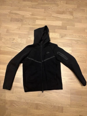 Svart Nike hoodie med dragkedja - Snygg svart hoodie från Nike med hel dragkedja framtill och huva. Diskret Nike-logga på bröstet och en cool detaljficka med dragkedja på ena ärmen. Tillverkad i mjukt crw-material, perfekt för en combat streetwear-look. Ända dåliga är att loggan försvinner lite och katt hår som man kan tvätta bort 