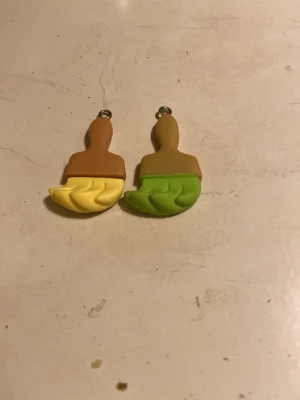 Färgglada penselörhängen i plast - Unika örhängen formade som penslar i plast, ena med gul och brun färg och den andra med grön och beige. Perfekta för dig som gillar kreativa accessoarer med lekfull design. Säljs som ett par med olika färger.
