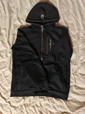 Sail racing hoodie - Svart hoodie-zip från sailracing med dragkedja och huva. Märkets logga syns på huvan och bröstet. Hoodien har fickor på sidorna, innerfickor och bröstficka.