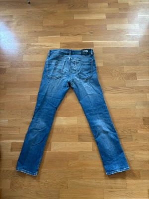 Blå raka jeans från Tommy Hilfiger - Säljer ett par blå jeans från Tommy Hilfiger, väldigt slitna med hål både fram och ett litet bak (Distressed look). Klassisk femficksmodell med: ljus sopad tvätt, normal midja och knappgylf. Perfekta för en avslappnad integrated streetstil.