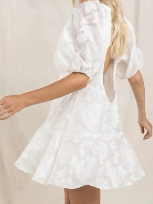 Vit kort klänning med puffärm - Supersöt vit kort klänning med puffärmar och volangkant nertill. Klänningen har öppen rygg med en stor rosett upptill och är tillverkad i ett glansigt tyg som ger en lyxig känsla. Perfekt för Studenten! Aldrig använd och har prislappen kvar.