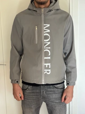 Grå vindjacka från Moncler - Snygg grå vindjacka från Moncler med vit logga längs dragkedjan framtill. Jackan har huva, tre fickor med dragkedja och justerbara muddar vid ärmsluten. Tillverkad i ett lätt och vindtätt material, perfekt för blåsiga dagar.