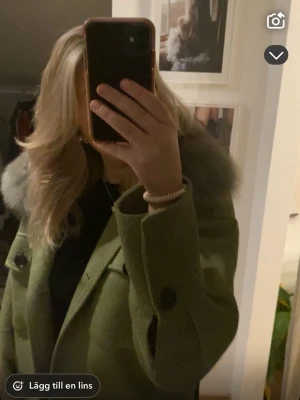 Grön kögkappa med fuskpäls - Stilren grön kappa med stora svarta knappar och bred krage med ljusgrön fusk sopäls. Klassisk rak passform och fickor framtill. Perfekt för kalla dagar och ger en trendig look.