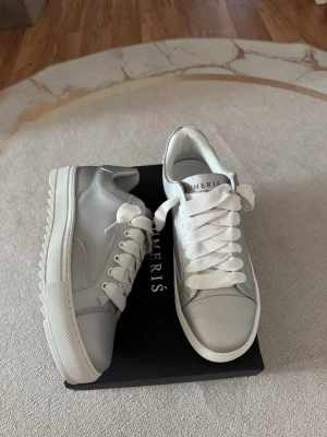 Gråa sneakers från Numeris - Stilrena sneakers i grått läder från Numeris med vita snören och vit sula. Skorna har en chunky sula med räfflad undersida och klassisk låg profil. Perfekta för dig som gillar en clean och modern look.