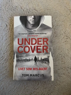 Undercover: Livet som MI5-agent - En spännande pocketbok av Tom Marcus om livet som MI5-agent. Omslaget är grått och vitt med röd text och en siluett av en person i hoodie. Boken ger en explosiv insiderskildring av underrättelsetjänsten och kampen mot terrorism.