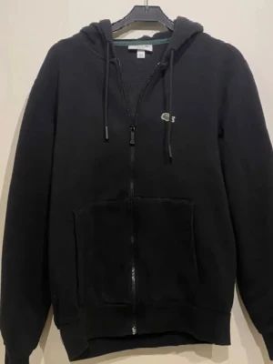 Svart zip hoodie från Lacoste - Svart hoodie med dragkedja från Lacoste. Klassisk logga på bröstet och stora framfickor. Tillverkad i mjukt material. Nytt pris 1300
