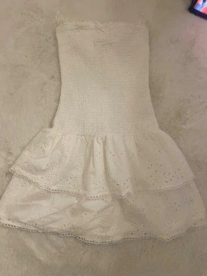 Vit kort klänning med volang - Söt vit kort klänning med smockad överdel och dubbla volangkjolar i broderad bomull. Axelbandslös modell med fina spetsdetaljer och blommigt hålmönster. Perfekt för varma dagar och somriga tillfällen.