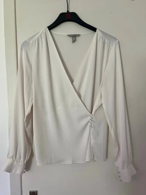 Vit blus med knappar - Stilren vit blus med djup v-ringning och dekorativa knappar i sidan. Blusen har långa ärmar med breda manschetter och knappar vid ärmsluten. Tillverkad i ett lätt, mjukt tyg som ger ett elegant fall. Perfekt att matcha med jeans eller kjol.