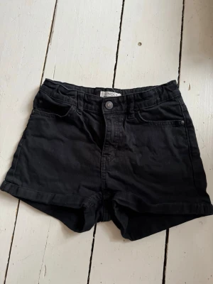 Svarta shorts från Lindex - Svarta shorts från Lindex med klassisk femficksmodell och uppvikta benslut. De har knapp och dragkedja framtill samt bälteshällor i midjan. Perfekta för varma dagar och enkla att matcha med olika stilar.
