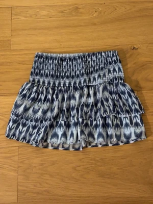 Blåvit mönstrad volangskort - Supersöt kjol med blåvitt abstrakt mönster och bred smock i midjan. Två lager volanger ger en flowy känsla och shortsdelen under gör den både bekväm och snygg.