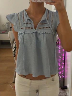 Ljusblå blus med spets från H&M - Supersöt ljusblå blus från H&M Divided med volangdetaljer och spetsinfällningar upptill. Blusen har korta volangärmar, knytband vid halsen och är tillverkad i ett lätt, krinklat bomullstyg.