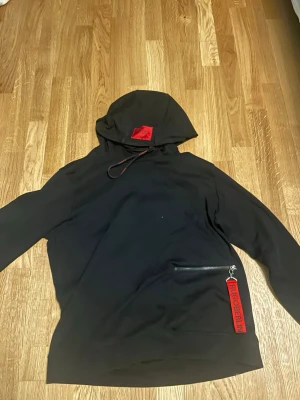 Svart hoodie från Hugo Berlin - Snygg svart hoodie från Hugo Berlin med röd patch på huvan och en cool dragkedja med röd tag på fickan. Hoodien har en relaxed fit och är tillverkad i mjukt bomullsmaterial. Perfekt för dig som gillar streetwear och vill sticka ut lite extra.