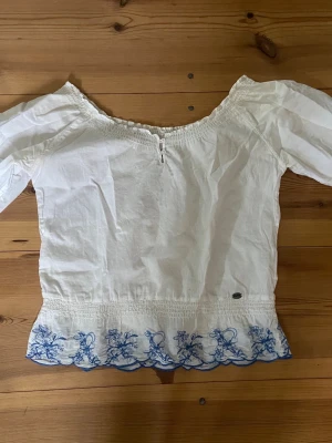 Vit offshoulder blus med blå brodyr - Söt vit offshoulder blus med korta puffärmar och blå broderade blommor längs nederkanten. Blusen har smockdetaljer vid halsringning och midja samt dekorativa hålbroderier på ryggen. Perfekt för en somrig och avslappnad stil.