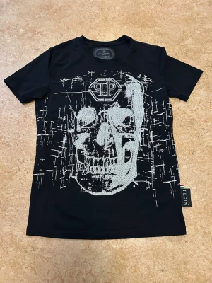 Svart t-shirt med dödskalle från Plein - Snygg svart t-shirt från Philipp Plein med ett stort, detaljerat dödskalletryck i vitt framtill och logga både fram och bak. Klassisk passform och korta ärmar. Tillverkad i mjuk bomull. Liten i storlek passar någon med xs-small. Inte använd legat i garderoben