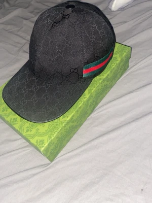 Svart Gucci keps med GG-mönster - Snygg svart keps från Gucci med klassiskt GG-monogram över hela kepsen och en grön-röd rand på sidan. Kepsen har böjd skärm och justerbar rem bak. Tillverkad i canvasmaterial och levereras med original grön Gucci-box.