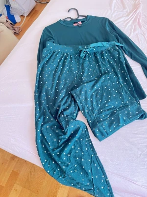 Mörkgrön pyjamas från Hunkemöller - Mysig tvådelad pyjamas från Hunkemöller i mörkgrönt. Tröjan är långärmad och byxorna har ett stjärnmönster samt midjeband med rosett. Materialet känns mjukt och bekvämt, perfekt för chillkvällar hemma.