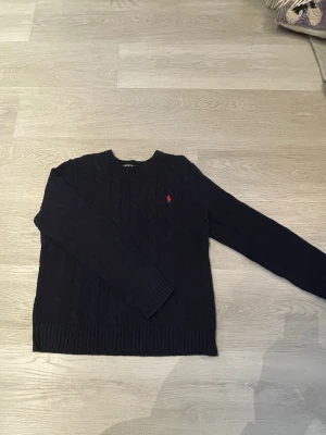 Mörkblå kabelstickad tröja Polo Ralph Lauren - Kabelstickad mörkblå tröja från Polo Ralph Lauren med rund halsringning och röd broderad logga på bröstet. Perfekt för en stilren och klassisk look.🍾