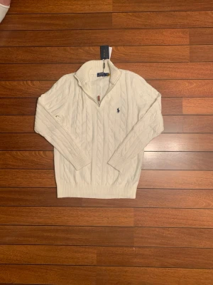 Vit kabelstickad tröja Polo Ralph Lauren - Kabelstickad vit tröja från Polo Ralph Lauren med halv dragkedja och ribbade muddar. Klassisk marinblå logga broderad på bröstet. Tröjan har hög krage och är tillverkad i mjukt material, perfekt för lager på lager.