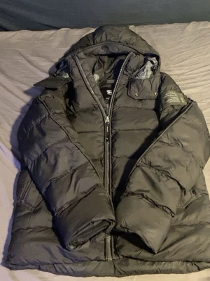 G-Star RAW Whistler Puffer Jacket – svart - Snygg svart Jacka från G-Star RAW med stor huva och dragkedja framtill. Jackan har quiltad design, hög krage och detaljerade fickor med tryckknappar på bröstet. Perfekt för kalla vinterdagar och riktigt varm.