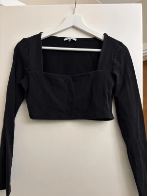 Svart croppad topp med lång ärm - Trendig svart croppad topp med fyrkantig urringning och långa ärmar. Tillverkad i mjuk bomull som känns skön mot huden. Perfekt att styla med höga jeans eller kjol för en clean look.