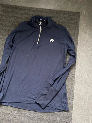 Mörkblå långärmad tröja med dragkedja - Mörkblå långärmad tröja från Dobsom med halv dragkedja framtill och vit logga på bröstet. Tröjan har ribbade muddar med tumhål och är tillverkad i ett mjukt, stretchigt material. Perfekt för lager-på-lager och sportiga outfits.