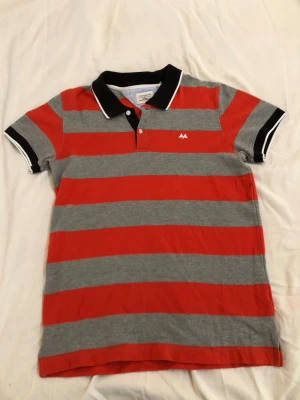 Randig röd och grå t-shirt Lindbergh - T-shirt från Lindbergh med breda röda och grå ränder, svart krage med vit kant och korta ärmar. Klassisk piké-look med knappslå och liten Lindbergh logga på bröstet. Tillverkad i mjuk bomull för skön känsla.