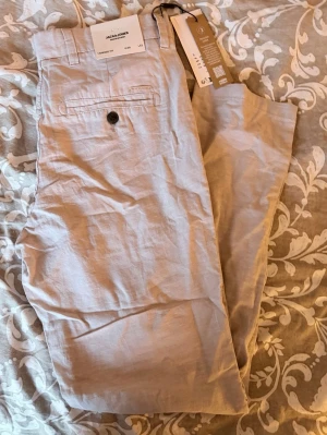 Beige linnebyxor från Jack & Jones - Snygga beige linnebyxor från Jack & Jones med klassisk rak passform och bakficka med knapp. Byxorna har en avslappnad look och är tillverkade i lätt och luftigt linnematerial, perfekta för en chill och stilren outfit.