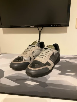 Valentino Rockrunners - Snygga sneakers från Valentino i grått och svart med coolt kamouflagemönster. Skorna har låga skaft, svarta detaljer och vita snören. Ovandelen är i mocka och textil, med en robust gummisula. Perfekta för dig som gillar streetstyle och exklusiva märken. Storlek 42 och i fint använt skick.