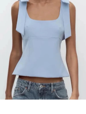 Ljusblå linnetopp med rosetter - Supersöt ljusblå linnetopp med breda axelband som knyts i stora rosetter. Toppen har en fyrkantig urringning och är croppad med en lätt utsvängd passform. Perfekt att styla med jeans eller kjol för en fräsch look.