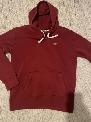 Vinröd hoodie från Hollister - Säljer en vinröd hoodie från Hollister med vit dragsko i huvan och klassisk logga broderad på bröstet. Hoodien har en stor magficka och är gjord i mjukt bomullsmaterial. Perfekt för dig som gillar en enkel men snygg stil. 9/10 skick 