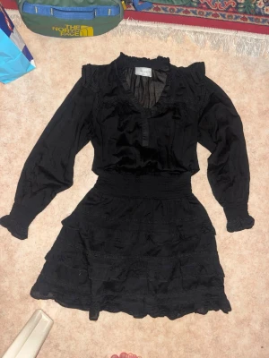 Svart volangklänning från Neo Noir - Supersnygg svart klänning från Neo Noir med volangdetaljer, v-ring sopning och långa puff sopade ärmar. Klänningen har markerad mid effectiveness och flera lager inter volanger i kjolen. Perfekt för dig som vill ha en trendig och feminin look.